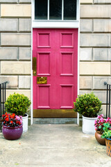 Brick residential building with a pink wooden door. Gilded metal details, iron gate and decorative plants and flowers at the entrance. Located in the New Town neighborhood of Edinburgh.
