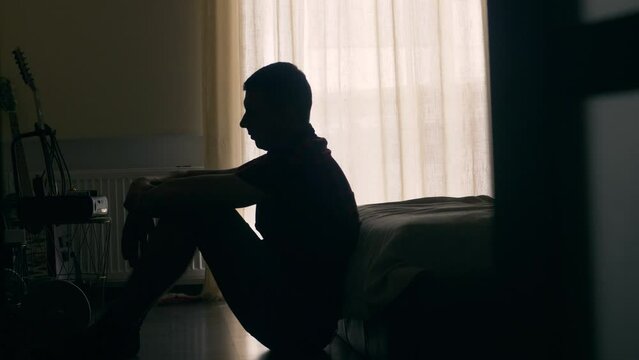 Silhouette of depressed man sitting on a floor. Sad unhappy person in a bedroom thinking about something