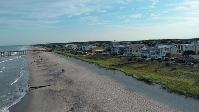 aerial footage along the Atlantic coastline at Kure Beach with people relaxing on the beach, blue ocean water, waves, green grass, trees and homes along the coast in Wilmington North Carolina USA