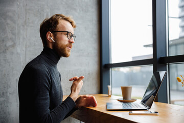 A young red-haired guy, a programmer or businessman in glasses, is sitting in a stylish cafe at a...