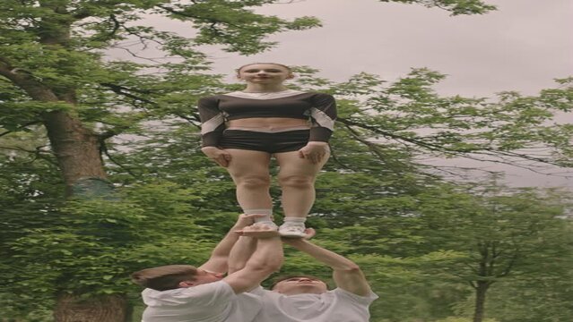 Vertical shot of top girl held by male athlete performing heel stretch during cheerleading performance on outdoor soccer field