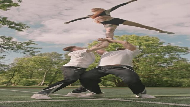Vertical shot of two base athletes holding flyer girl above heads and she doing arabesque pose while practicing cheerleading extension stunt outdoors on sports field