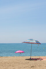 dos sombrillas plantadas en la arena al borde del mar con un velero en el horizonte y un cielo azul.