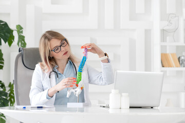 Young attractive female doctor orthopedist demonstrating the problem on spine bone model on the desk in her workplace