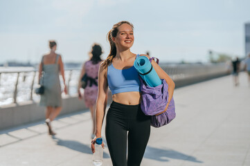 Beautiful fit caucasian girl in sportswear walks at embankment carrying yoga mat bottle of water...