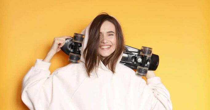 Stylish young happy girl in white hoody smiling while holding skateboard on shoulder isolated on yellow background. Enjoying life. Healthy and careless lifestyle.