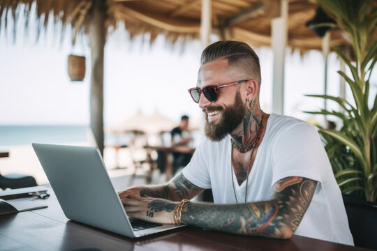 Smiling handsome hipster man with tattoos sitting and working on his laptop in the beach bar