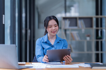 Fototapeta premium Smiling female browsing laptop and tablet sitting at table with papers and books while in the office.