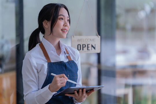 Asian business woman in an apron, the owner of the cafe stands at the door with a sign Open waiting for customers , cafes and restaurants Small business concept.