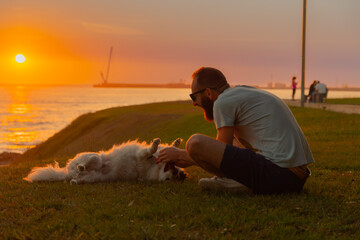mature 40s man playing with dog at sunset sitting grass watching the sea and the porto lighthouse,...