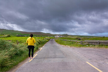 Fototapeta premium Lonely person with yellow jacket standing on the road through green fields on a cloudy stormy day