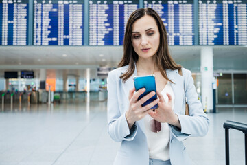 business woman wearing a suit and holding a phone standing at an airport in front of a display...
