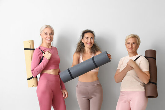Sporty mature women with yoga mats near light wall