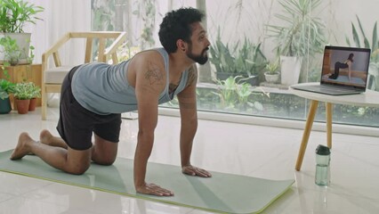 Man doing cat and cow exercise on fitness mat and watching video workout on laptop while practicing yoga at home