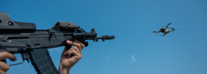 A man aims to shoot a rifle at a flying drone against a blue sky. 