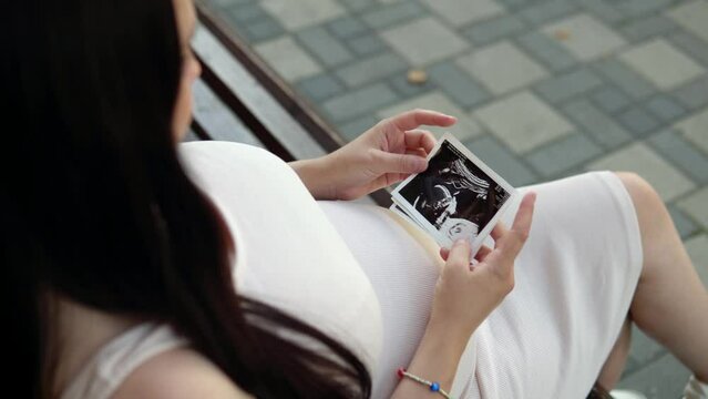 Amidst the crowd, a pregnant woman finds a quiet moment on a park bench, studying an ultrasound image of her child.