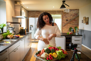 Middle aged female mixed ethnicity food nutritionist and blogger recording a video about a healthy recipe with organic fruits and vegetables in her kitchen at home