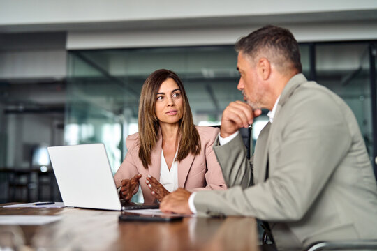 Busy Colleagues Executives Using Laptop Discussing Digital Project Commercial Tech Solution At Meeting Table. Two Professional Mid Aged Business Man And Woman, Manager And Client Talking In Office.