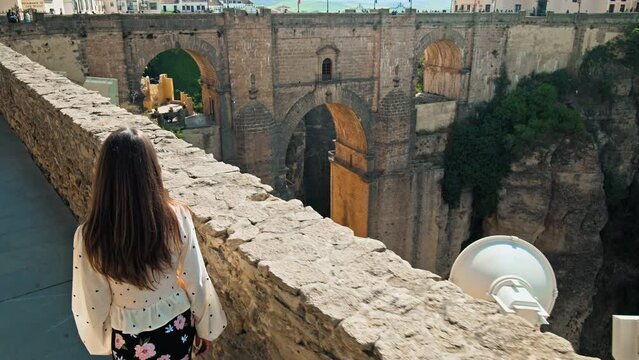 A girl tourist walking by the New Bridge Interpretation Center in Ronda, Spain. A female exploring looking at the Centro de Interpretaci&oacute;n del Puente Nuevo with a huge medieval arch bridge