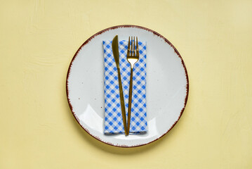 Empty plate with fork, knife and blue checkered napkin on yellow background