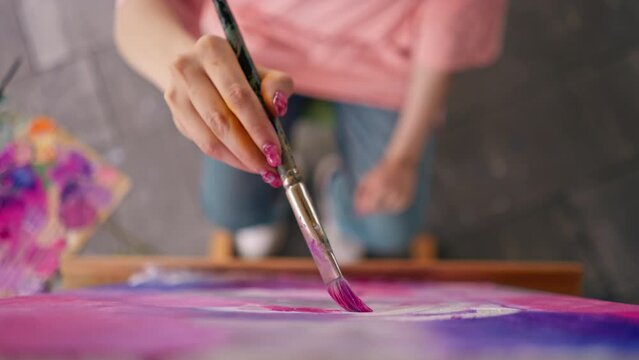Close-up of a girl artist with a brush applying oil paint to a painting standing on an easel in a studio for painting 