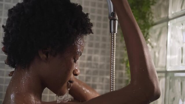Young black afro female taking relaxing shower at home. Adult African American woman gets hair and head wet enjoying self-care in bathroom. Concept of daily domestic personal hygiene in morning.