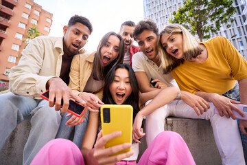 Group young people surprised watching something on phone. Multiracial friends sitting outdoor using...