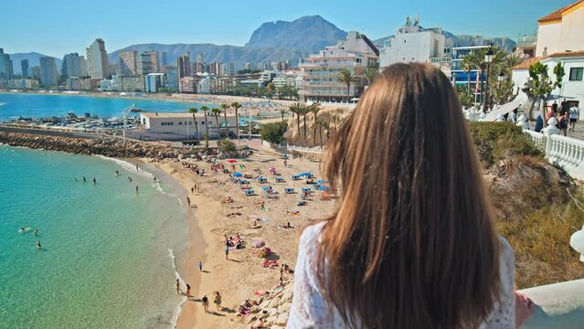 A beautiful girl in a dress looking at a scenic view of Spanish Beach and the Sea. A woman enjoying panoramic views of Platja de Llevant and Cala del Mal Pas Beach from above in Benidorm, Alicante