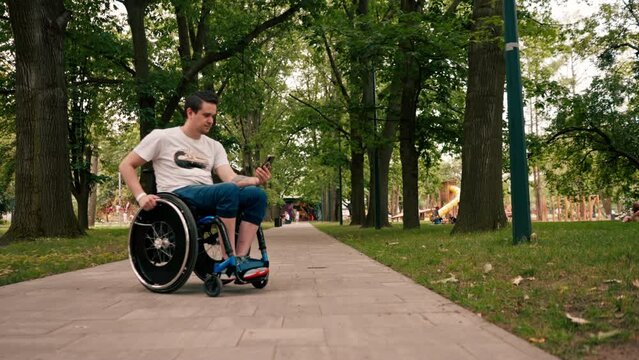 young man in a wheelchair looks intently at the phone emotionally reads the news or communicates in the messenger in the city park technology mobility