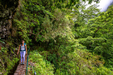 Fototapeta na wymiar Backpacker tourist walks along picturesque hike trail overgrown by plants through Madeira rainforest. Levada of Caldeirão Verde, Madeira Island, Portugal, Europe.