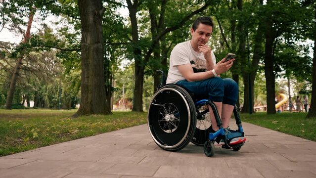 young man in a wheelchair looks intently at the phone emotionally reads the news or communicates in the messenger in the city park technology mobility