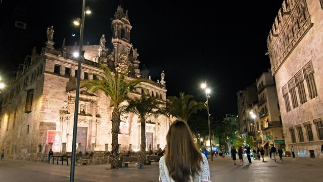 Girl tourist walking by Real Parroquia de los Santos Juanes Valencia at night. A tourist woman exploring a Long-standing Catholic church featuring a triangular bell tower topped with statues in Spain