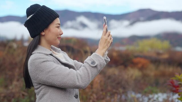 Asian woman using mobile phone taking selfie or vlogging with mt Fuji covered in snow with beautiful red maple tree leaves falling during travel nature lake Kawaguchi, Japan in autumn holiday vacation