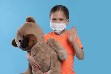 Cute girl with medical mask and teddy bear on light blue background