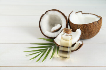 Bottle of organic coconut cooking oil, leaf and fresh fruits on white wooden table, space for text
