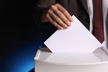 Man putting his vote into ballot box on dark blue background, closeup. Space for text