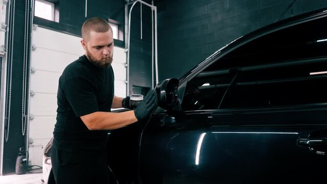 A Focused Male car wash worker in black gloves polishes the mirror of a luxury blue car using polishing sponge car care concept