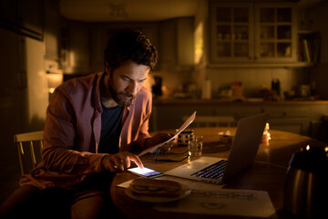 Young caucasian man calculating his bills on a smartphone in the kitchen