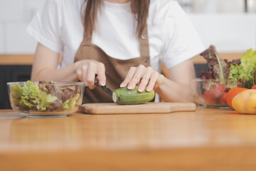 Delicious fruit and vegetables on a table and woman cooking. Housewife is cutting green cucumbers on a wooden board for making fresh salad in the kitchen.