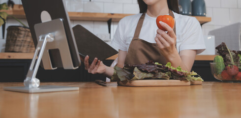 Delicious fruit and vegetables on a table and woman cooking. Housewife is cutting green cucumbers on a wooden board for making fresh salad in the kitchen.