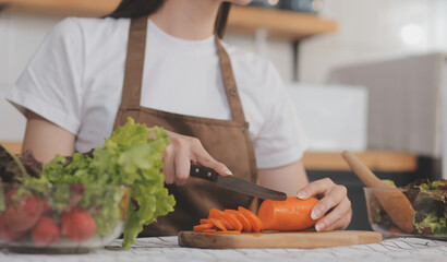 Delicious fruit and vegetables on a table and woman cooking. Housewife is cutting green cucumbers on a wooden board for making fresh salad in the kitchen.