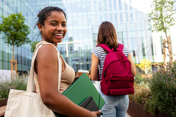 Happy Black female college student standing outside university building holding notebooks in campus...