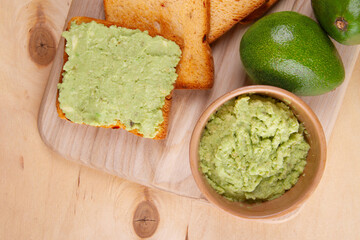 On pale yellow wooden surface you'll find still life with avocado toast, luscious avocado spread and two perfectly fresh avocado fruits close-up