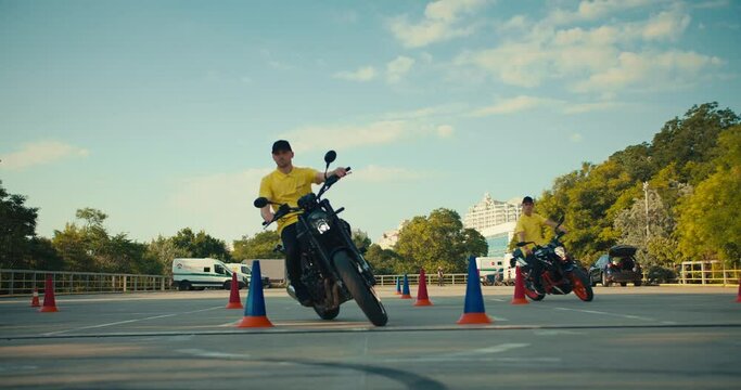 Two driving instructors in yellow t-shirts hone their skills in motorcycle maneuverability. Two guys go around obstacles on a motorcycle