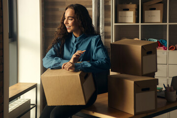 A young woman sits on a table in front of boxes, holding them in her hands, collecting parcels at the post office. entrepreneur online store