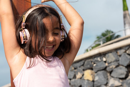 Girl with bright pink headphones and dressed in pink, with short hair. She is smiling, listening to music in a park with her hands up dancing, having a fun time