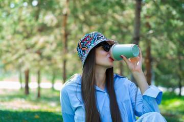 Summer portrait of a girl with long blond hair in sunglasses. A girl in a blue shirt and blue jeans and a panama sits on the grass and drinks coffee from a thermo mug.