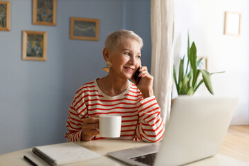 Cute stylish senior smiling female freelancer in striped shirt drinking tea and talking on phone...