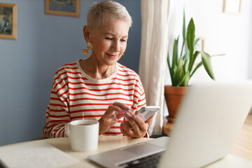 Selective focus of good-looking female of 60s with stylish hairstyle checking online messenger, chatting in dating app or browsing social media sitting at table in front of laptop and cup of tea