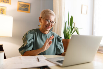 Senior trendy elegant female teacher in glasses with stylish haircut waving hand at camera of laptop saluting her students in beginning of online lesson, sitting at table of her home cabinet